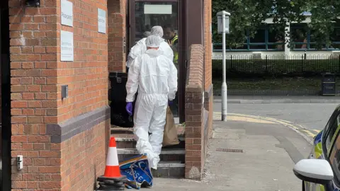 Two people wearing white forensic suits and purple gloves entering a brick building on Bedford Street. One of them is carrying a brown paper bag as he walks up the stairs into the property. A police car can be seen parked to the right.