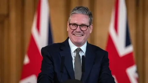 Sir Keir Starmer standing in the Number 10 briefing room. He is smiling at the crowd. He wears a dark suit, white shirt, grey tie and black-rimmed glasses. Behind him are two union jack flags.