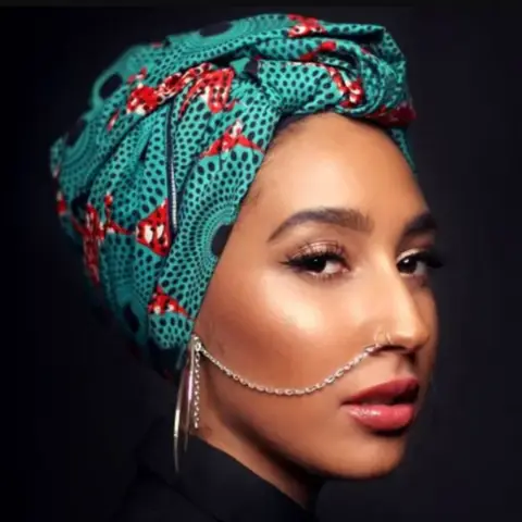 INSANITY PR Mariah is wearing a green and orange headscarf with a nose chain extending to her ear. She has make-up on and is looking at the camera. 
