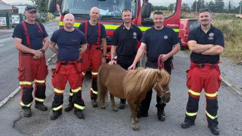 Kieran Moyes A group of male firefighters stand in front of a fire engine alongside a small, brown pony. 