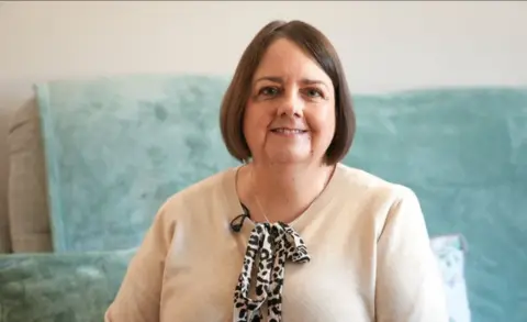 BBC Ceri Maher is smiling at the camera. She has a cream-coloured jumper on with a leopard-print tie at the front. Her brunette hair is in the shape of a bob cut and behind her is a cream sofa that is covered by a mint-green throw. 