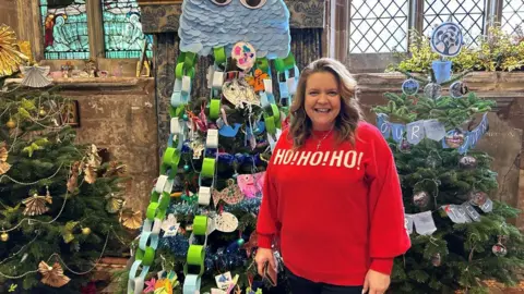 A smiling woman with blonde hair and a bright red jumper with "ho ho ho" in white lettering in front of three decorated Christmas trees and a number of large church windows