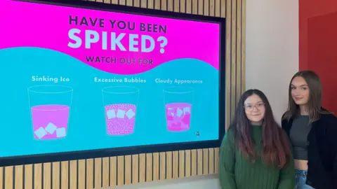 Two women, one with long brown hair, glasses and a green jumper, one with brown hair cropped top and cardigan, standing next to a pink 'Have you been spiked poster.'