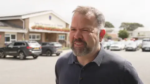 A middle aged man is pictured in a car park. He is wearing a black polo shirt, has short brown/grey hair and a stubble beard. He is smiling.