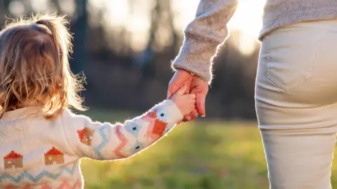 Getty Images A rear view shot of an unidentified toddler with long hair and a cardigan holding hands with an unidentified adult in cream jeans and a grey jumper. 