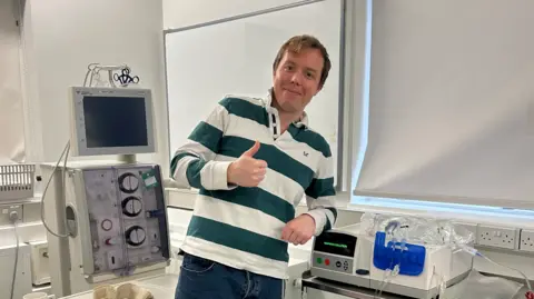 Cameron Frazer A man with brown hair and a striped green and white long sleeved tops leaning on a white piece of medical equipment with a thumbs up and with other medical equipment behind him