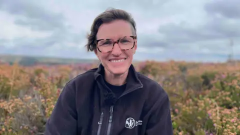 Joanna Hill, wearing a black zip-up fleece and patterned-rimmed glasses, smiling with heather in the background and a cloudy sky.