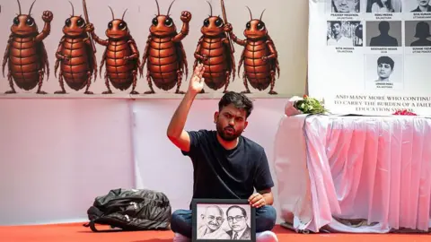 Getty Images Cockroach Janata Party founder Abhijeet Dipke wears a black tee shirt as he sits cross legged on a carpet at the protest side, holding a portrait of M K Gandhi and Independent India's first law minister B R Ambedkar. Behing him, there's a flex board with animated pictures of six cockroaches and another board with photographs of students who reportedly died by suicide after the recent medical entrance exam cancellation die to paper leak.