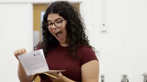 A girl with long curly dark hair and glasses, with her mouth open in exclamation as she reads a letter.