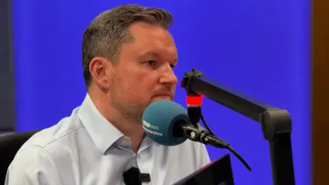 Kevin McNamara, a man with greying hair, sits in front of a branded microphone in the BBC Radio Gloucestershire studio. There is a purple lightbox behind him. He is wearing a pale blue shirt.
