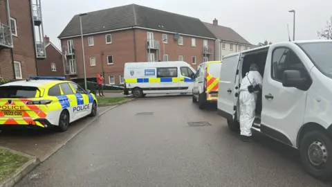 Nicola Haseler/BBC A police car and two police vans along with a white forensic van are parked on a residential street in front of a police cordon. Blocks of flats are pictured in the distance. A forensic officer can be seen looking into the site of the white van.