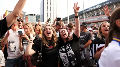 Reuters A large crowd of people gathered outside the Principality Stadium in Cardiff. Many are wearing black T-shirts featuring the band Oasis, and some are holding drinks or mobile phones aloft. Several individuals have their arms raised in the air, creating a lively and celebratory atmosphere. In the background, modern buildings and the stadium’s signage are visible under a bright, clear sky.