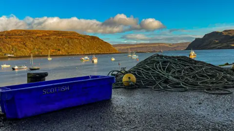 JESSE LEITE An empty blue box with the word Scottish on the side sat next to a pile of ropes on the side of a harbour with fishing boats in the background. 