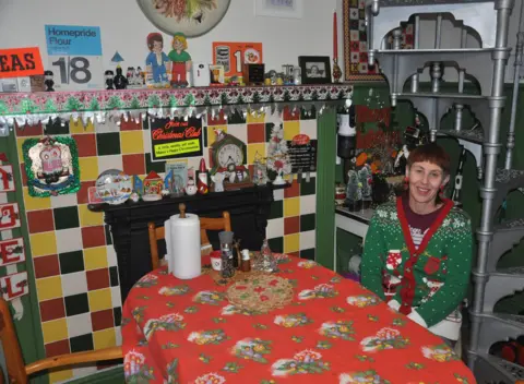 Lisa Watts sits at a small kitchen table covered with a red tablecloth, surrounded by colourful tiled walls, vintage Christmas decorations on the mantel, and shelves filled with festive ornaments.