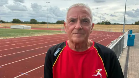 Ian Richards - a man with short, white hair - is looking into the camera and wearing a red and black athletics top. A red running track can be seen behind him.