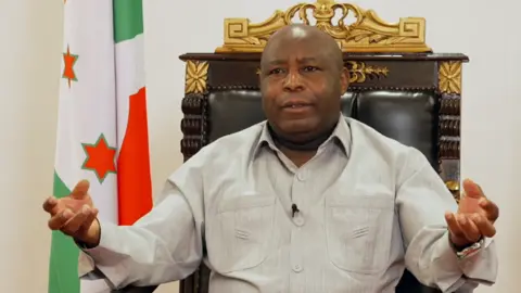 President Évariste Ndayishimiye sits on a wooded and gilt chair, next to the Burundian flag. He is gesticulating with his hands while talking.