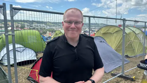 A police officer wearing a black, short-sleeved top and glasses smiles at the camera. He is standing in front of a metal fence with tents behind hit.