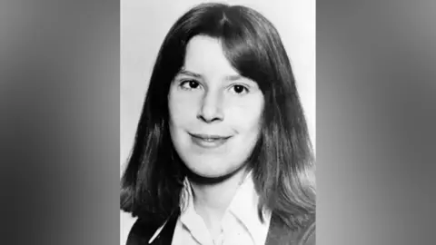 A black and white image of Suzanne Lawrence, who has shoulder-length hair and wears a shirt and cardigan. 