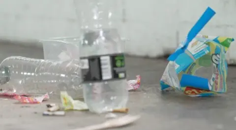A close-up image of rubbish on a car park floor. It includes water bottles, sweet packets, wrappers and a plastic tub.