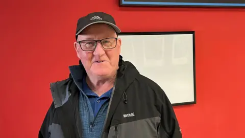 Jonny Humphries/BBC Tony Finnegan, 66, who is clean-shaven and is wearing glasses, a black Adidas branded baseball cap and a black and grey raincoat over a blue polo-neck t-shirt, smiles at the camera standing in front of a painted red wall.  