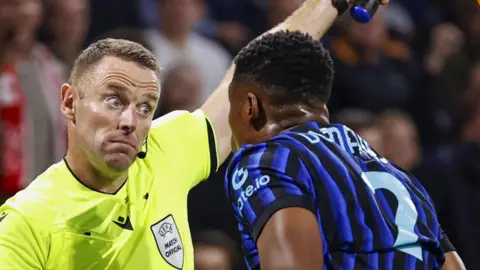 A man in a luminous green shirt holds up a yellow and orange chequered flag in his left hand while pointing his right hand at a footballer in a blue and black striped football top. A player in a red and white football top walks past. Spectators are in the background.
