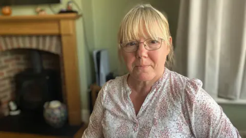 BBC Julie Ray, wearing glasses with her blonde hair up, sits in her front room in Didcot and looks at the camera