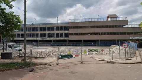 Metal fences surround the perimeter of a demolition site in Paignton. Large blocks of rubble are on the floor and a white truck is parked on the site. A multi-storey car park is in the background.