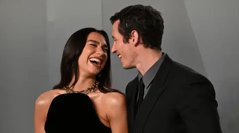 PA Media Red-carpet photograph of Dua Lipa and Callum Turner. Lipa is wearing a black dress with a large gold necklace, and has long dark hair. She is smiling, and looking up at Turner, who is wearing a black suit, a grey shirt, and a black tie. The couple are stood in front of a dark grey backdrop. 