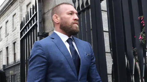 PA Two men walk out a black gate, one behind the other. The man at the front is wearing a navy suit, navy tie and white shirt, he has ginger short hair and a ginger beard.
The man behind him wears a black suit and tie with a light blue shirt.