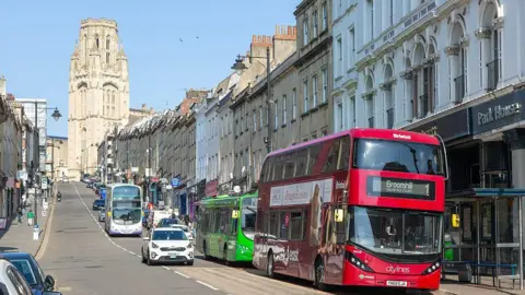 Getty Images Three buses driving or parked on Park Street in Bristol. The street is a long steep hill with a large, ornate tower at the top end. 