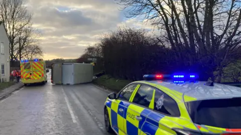 Paul Pinkstone Police officers and recovery crews attend a road incident on a rural route, with police vehicles blocking the carriageway and a recovery truck at the scene.