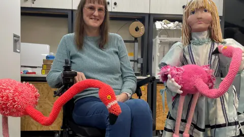 Rugby Yarn Bombers A woman wearing glasses, jeans and a jumper smiles at the camera. She is in a wheelchair and has a crafted orange flamingo next to her. Beside the woman is a knitted Alice who is holding a crafted flamingo.