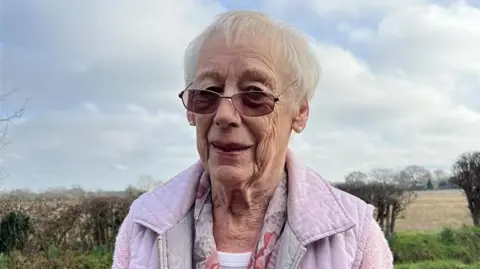 Barbara Kamp is outside with fields and a hedgerow behind her. She is wearing sunglasses and a pink gilet with a pink jumper and a patterned scarf. She has short white hair.