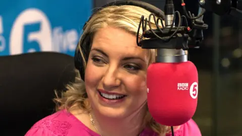 BBC Anna Foster pictured in front of a microphone in the BBC Radio 5 Live studio. She is wearing a pink top.