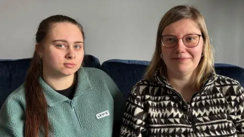 On the left, there is a girl with a turquoise zip-up hoodie, with "Levi's" written in black capital letters on a white background, on it. She also has brown long hair, going over her shoulder, and a black t-shirt. On the right, there is a woman with black thin-framed glasses, blonde hair over her shoulders, and a black and white zip-up hoodie.