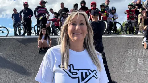 Simon Thake/BBC A blonde woman with shoulder-length hair smiles. She is wearing a bright white T-shirt. Behind her young BMX riders sit at the top of a ramp.