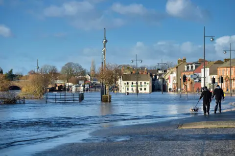 Getty Images The River Nith spills out across the Whitesands covering the road and nearby car park. Two people are out walking a dog next to the lapping water.