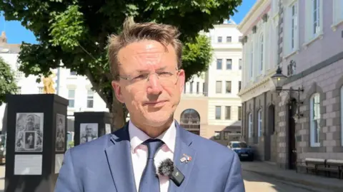 Jersey minister Deputy Kirsten Morel. He is wearing a blue suit and blue tie. He is standing in front of white stone buildings.
