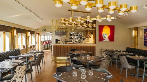 Jack Spicer Adams The interior of a pub, featuring dozens of tables and chairs. Glasses and cutlery lie on the tables. A bar can be seen in the background, alongside a bright portrait hanging on the wall.