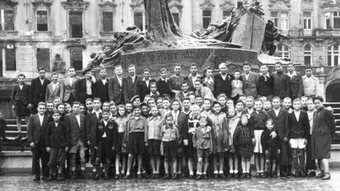 BBC/Wall to Wall/Lake District Holocaust Project A black and white picture of a group of children in Prague before they were brought to the Lake District. They are standing in front of a large statue or monument.