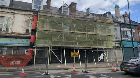 BBC/Mick Lunney Scaffolding surrounds the front of a partially collapsed building. The scaffolding is covered in a yellow mesh.