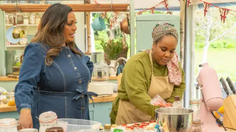 Channel 4/Love Productions/Mark Bourdillon Alison Hammond dans une robe en jean aux cheveux bruns bouclés se tient à gauche. Illyin Morrison prépare du glaçage rouge. Elle porte un pull vert et un tablier de boulanger. 