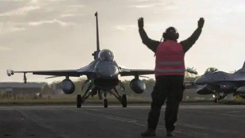 Royal Air Force A sleek military jet taxis on the RAF Waddington runway for Exercise Cobra Warrior. A member of the ground crew, wearing a pink hi-vis vest, holds his hands in the air as he signals to the pilot.