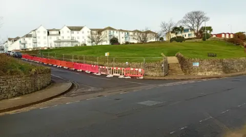 Dorset Coast Forum Green area surrounded by a stone wall and road running alongside, red and white barriers are in place along one side of the wall.