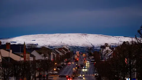 Pascal Diamond A white capped snowy hill sits above Cookstown main street, its evening and the road is busy with cars and buses