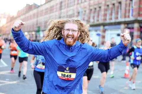 Bearded runner with long curly hair and spectacles celebratrs crossing the finish line in 2023 half marathon.