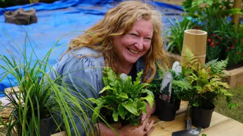 Chorley Council Charlie Dimmock, with long wavy ginger hair leans over a table in the garden grinning and hugging a plant. There are other plants on the table and in the background is a blue tarpaulin covering part of the garden