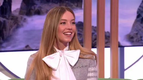 Sydney Sweeney smiles in The One Show studio, wearing a large white bowtie and a grey cardigan. She has long brown hair