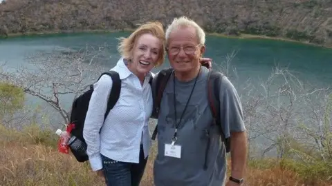 Royal Papworth Hospital Charity Wendy J. Tomlin-Hess is on the left. She has short dark blonde hair, and is wearing a blue shirt, dark blue jeans and a black rucksack. On the right is Robert Hess who has short grey hair and is wearing glasses, a blue T-shirt with a bull on it, a lanyard and a black rucksack. 