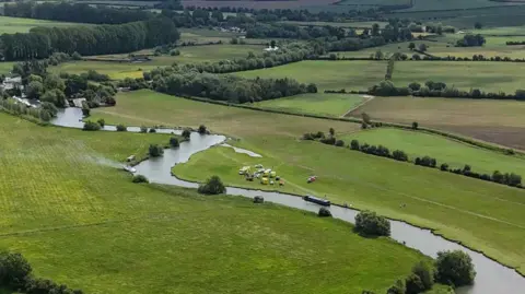 An aerial image showing the houseboat fire in Lechlade on Thames. The river meanders through vast green fields. On the left there is white smoke coming from a boat in the water. Parked on the right side of the river there are multiple emergency vehicles and a red helicopter.